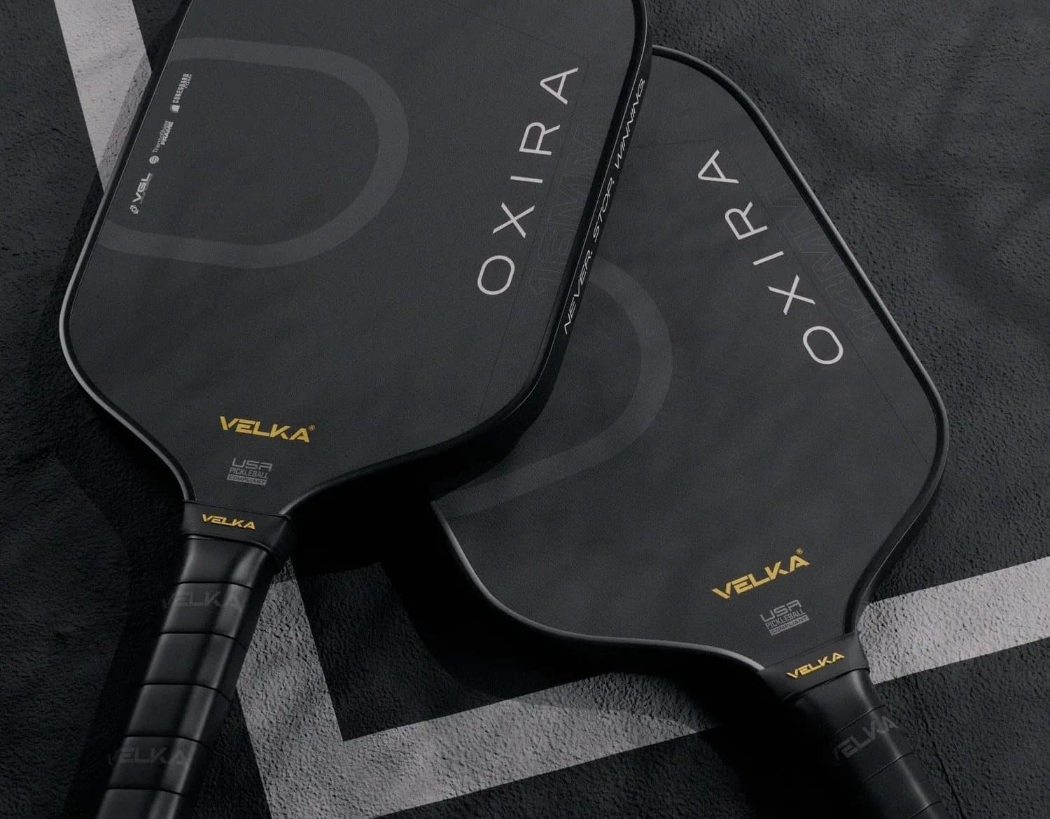 Two Velka Oxira pickleball paddles lying face up on a dark court beside the service line